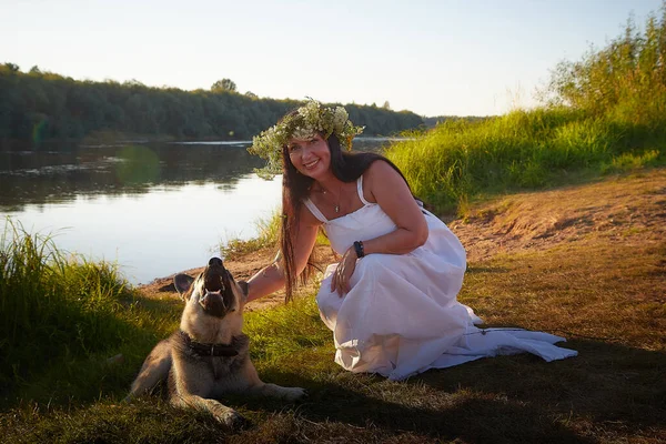 Adult mature brunette woman in white dress, sundress and wreath of flowers with big shepherd dog in summer by the water of river or lake in evening at sunset celebrating pagan holiday of Ivan Kupala