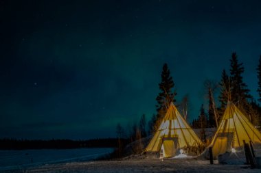 Kuzey Işıkları (Aurora Borealis), Kanada 'nın Kuzeybatı Bölgesi, Yellowknife yakınlarındaki ormanlarda aydınlatılmış çadırlar.
