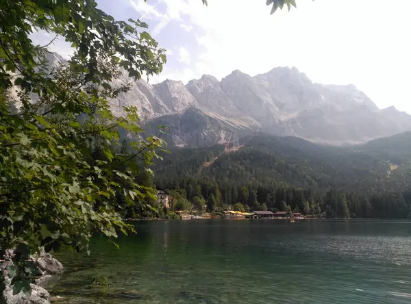 Ein wunderschoener Blick auf einen See und auf die Berge. Ben Vordergrund kann man einen Baum erkennen im Hintergrund ein Dorf, das genau vor den Bergen sitzt. Die Berge erstrecken sich in den klaren blauen ettd Wolkigen Himmel.