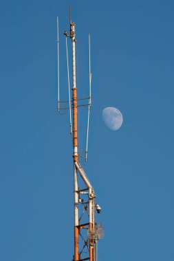 Yüksek bir iletişim anteni ve açık mavi gökyüzünde gündüz vakti bir ay..