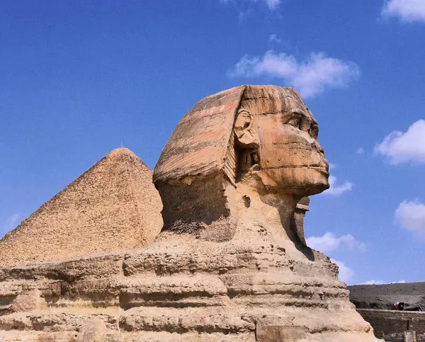 stock image Profile view of the Great Sphinx of Giza, with one of the Pyramids behind and a blue sky and wispy clouds in background in dynamic high contrast colour