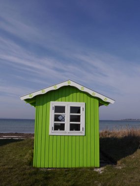 Beyaz çatısı ve pencere çerçevesi mavi deniz ve mavi gökyüzünün arka planında şirin, ıhlamur yeşili bir sahil kulübesi..