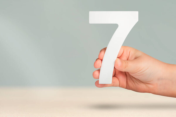 Number seven in hand. Hand holding white number 7 on blurred background with copy space. Concept with number seven. Birthday 7 years, percent, seventh grade or day