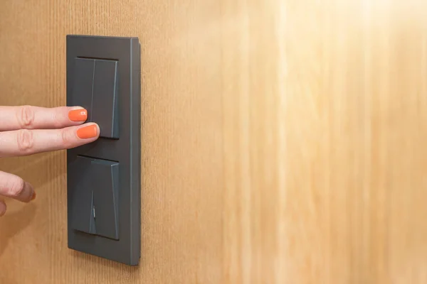 Turn on the light, a womans hand presses the switch in the apartment close-up. Black light switch on the background of a wooden wall. The concept of saving electricity, the cost of electricity