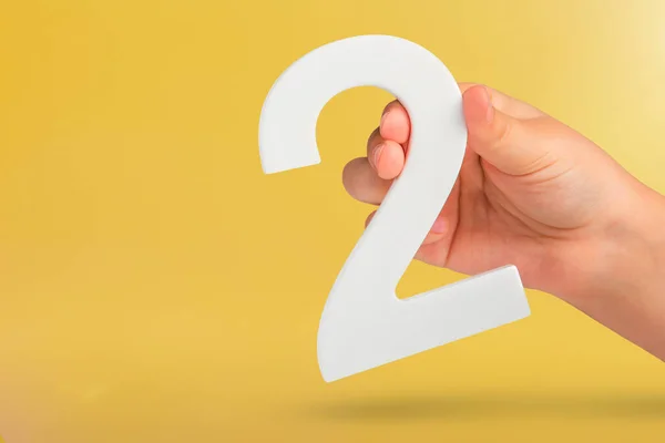 Number two in hand. Hand holding white number 2 on yellow background with copy space. Concept with number two. 2 percent, birthday 2 years old, couple, two, double.