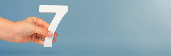 Номер семь в руке. Рука держит белый номер 7 на синем фоне с копировальным пространством. Концепция с номером семь. День рождения 7 лет, процентов, седьмой класс или день