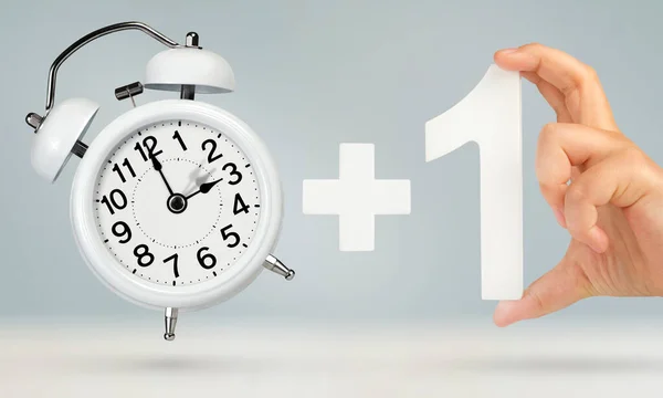 Change time. Spring forward, on a gray background. A white alarm clock indicates that the time has been moved forward an hour. Hand holding number one with plus sign, Spring forward concept.