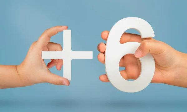 The number six and the plus symbol in the hands of a child on a blue background. White number 6 with a plus close-up. The concept of addition or sum of the number six.