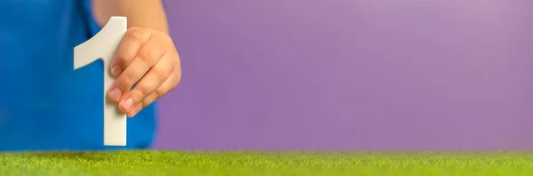Banner with hand holding number one on green grass and purple background. A symbol of unity, uniqueness and beginning, joining forces to achieve a common goal, the first step in a certain direction