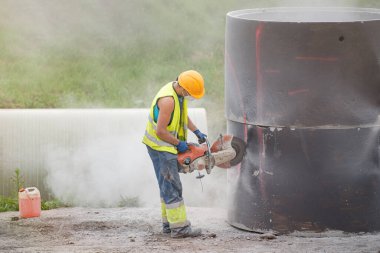 Yağmur suyu toplamak için yere beton boru döşüyorlar. İşçi, kanalizasyona bağlanmak için borunun beton kısmında delikler açıyor. İnşaat kıyafetleri içinde bir adam beton kesiyor.
