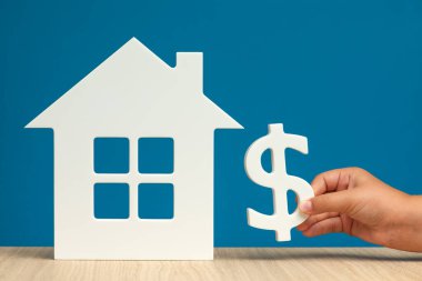 Amerika 'da emlak kiralamanın maliyeti. Amerika 'da emlak kiralamak. Beyaz bir ev modeli ve yakın plan mavi bir arka plan üzerinde dolar sembolü olan bir el. Boşluğu kopyala.