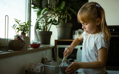 Modern bir mutfakta annesinin ev yapımı hamur işi yapmasına yardım eden tatlı küçük bir kız. Çocuk, metal kasede malzemeleri karıştırıp evde yemek pişirirken eğleniyor..