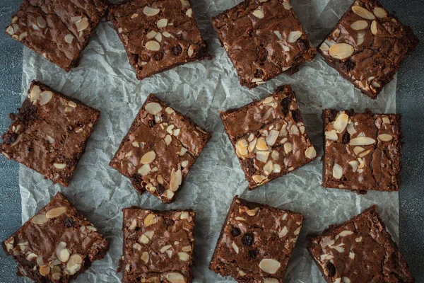 stock image Delicious homemade chocolate brownies with almond flakes and raisins cut on square slices on the baking parchment. Flat lay. Grey concrete background