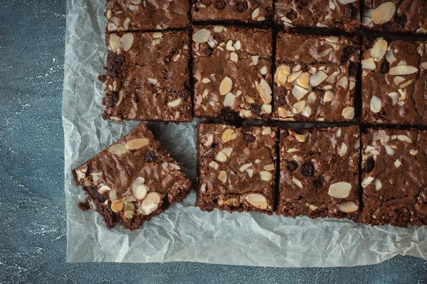 stock image Delicious homemade chocolate brownies with almond flakes and raisins cut on square slices on the baking parchment. Flat lay. Grey concrete background