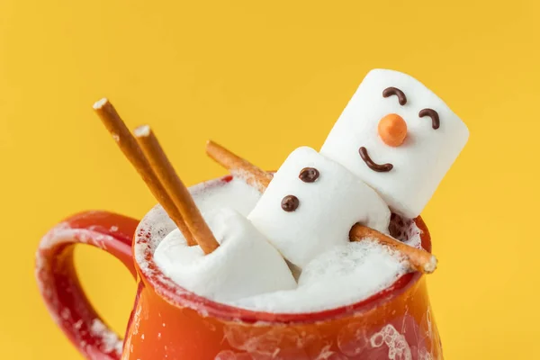 Marshmallow snowman taking hot tub in a red ceramic cup full of cocoa with milk foam. Christmas holidays yellow background. Wintertime concept. Hot chocolate with marshmallow and festive decoration.