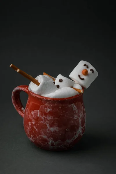 Marshmallow snowman taking hot tub in a red ceramic cup full of cocoa with milk foam. Christmas holidays grey background. Wintertime concept. Hot chocolate with marshmallow and festive decoration.