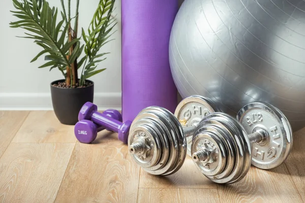 Light and heavy steel dumbbells, fitness ball and other sports equipment on the wooden floor in the gym. Fitness gear in the home interior. Training at home concept