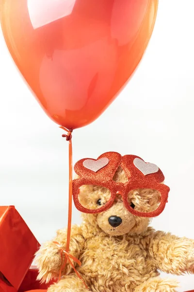 Little fluffy teddy bear wearing red heart shaped glasses and holding red balloon. Happy Valentine\'s Day. Presents in a red wrapping paper with golden bow isolated on the white background.