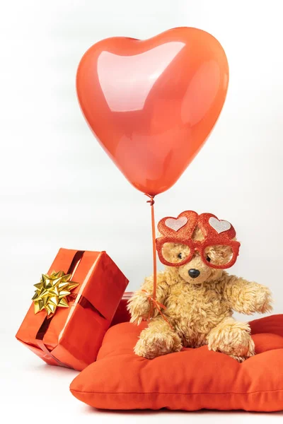 Little fluffy teddy bear wearing red heart shaped glasses and holding red balloon. Happy Valentine\'s Day. Presents in a red wrapping paper with golden bow isolated on the white background.