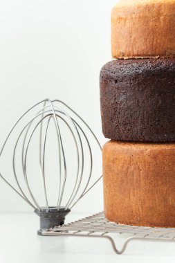 Freshly baked sponge biscuit on a pastry grid next to whisk, sieve for sifting flour and eggs. Brown chocolate and white vanilla sponge cake with professional confectioner utensils. White background