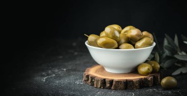 Close up shot of green olives in a white bowl on the wooden stand with olive leaves on a black background. Traditional Greek and Italian food.