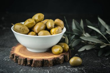 Close up shot of green olives in a white bowl on the wooden stand with olive leaves on a black background. Traditional Greek and Italian food.