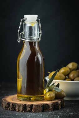 Green olives in a white bowl next to a bottle with olive oil and leaves on a black background. Bottle of cold pressed oil. Traditional Greek and Italian food.