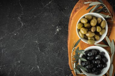 Green and black olives in white bowls on the wooden board and leaves on a black background. Traditional Greek and Italian food. Flat lay. Copy space