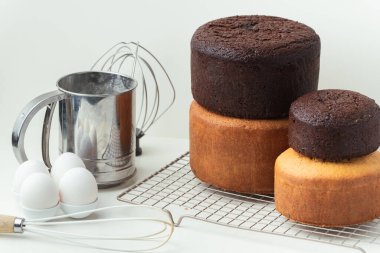 Freshly baked sponge biscuit on a pastry grid next to whisk, sieve for sifting flour and eggs. Brown chocolate and white vanilla sponge cake with professional confectioner utensils. White background