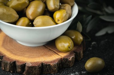 Close up shot of green olives in a white bowl on the wooden stand with olive leaves on a black background. Traditional Greek and Italian food.