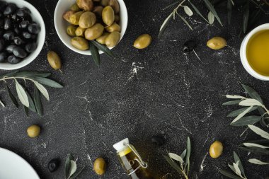 Green and black olives in white bowls next to a bottle with olive oil and leaves on a black background.