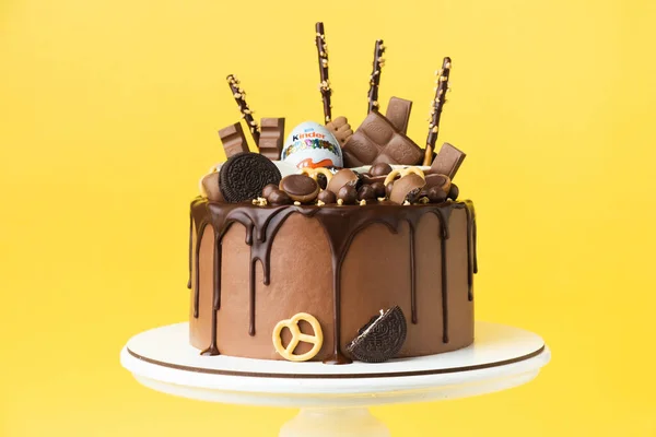 KYIV, UKRAINE - January 15: Birthday cake with brown cream cheese frosting and chocolate drips decorated with Kinder egg toy, Milka and KitKat sweets, Oreo cookies and candies. Yellow background