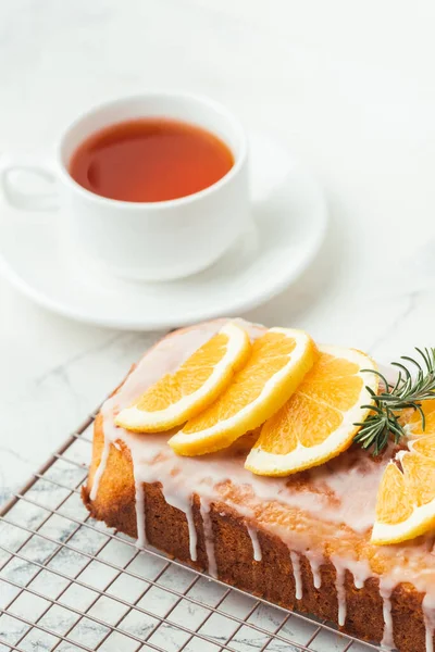 Loaf of orange bread covered with a confectionery glaze with lemon juice and decorated with orange slices. Chiffon cake on a pastry grill next to cup of tea. White background