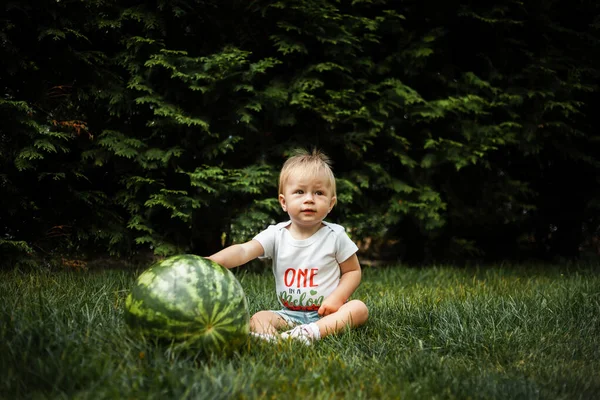One year old caucasian baby girl with blonde hair sitting on the grass next to big watermelon. Cute little child smiling in the park against green trees. Summer time concept