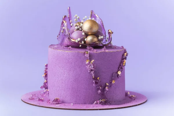 Wedding cake with violet cream cheese frosting decorated with caramel vase and golden chocolate spheres on the purple background. Luxury anniversary cake with pink chocolate velvet coating