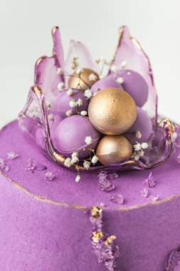 Wedding cake with violet cream cheese frosting decorated with caramel vase and golden chocolate spheres on the white background. Luxury anniversary cake with pink chocolate velvet coating