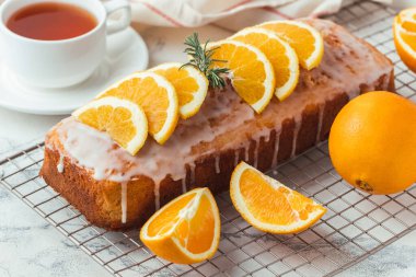 Loaf of orange bread covered with a confectionery glaze with lemon juice and decorated with orange slices. Chiffon cake on a pastry grill next to cup of tea. White background