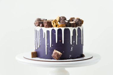 Birthday cake with dark blue cream cheese frosting with white chocolate drips on the white background. Homemade cake decorated with chocolate sweets and cookies on the white stand