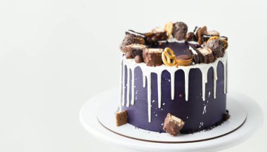 Birthday cake with dark blue cream cheese frosting with white chocolate drips on the white background. Homemade cake decorated with chocolate sweets and cookies on the white stand