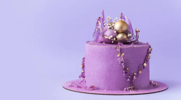 Wedding cake with violet cream cheese frosting decorated with caramel vase and golden chocolate spheres on the purple background. Luxury anniversary cake with pink chocolate velvet coating