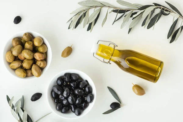 Green and black olives in white bowls next to a bottle with olive oil and leaves on a white background.