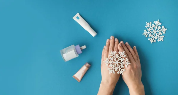 Using moisturizing hand cream during winter season. Female hands smearing white cream on the blue background next to different skin care products. Treatment for the skin advertisement