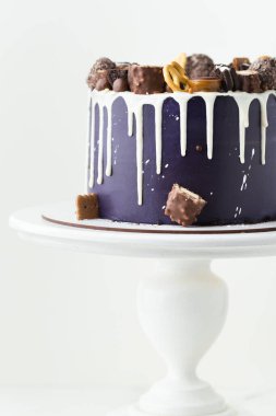 Birthday cake with dark blue cream cheese frosting with white chocolate drips on the white background. Homemade cake decorated with chocolate sweets and cookies on the white stand