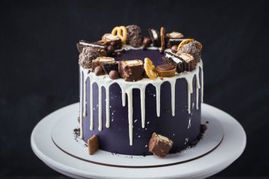 Birthday cake with dark blue cream cheese frosting with white chocolate drips on the black background. Homemade cake decorated with chocolate sweets and cookies on the white stand