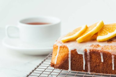 Loaf of orange bread covered with a confectionery glaze with lemon juice and decorated with orange slices. Chiffon cake on a pastry grill next to cup of tea. White background