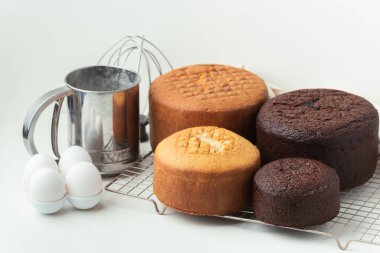 Freshly baked sponge biscuit on a pastry grid next to whisk, sieve for sifting flour and eggs. Brown chocolate and white vanilla sponge cake with professional confectioner utensils. White background