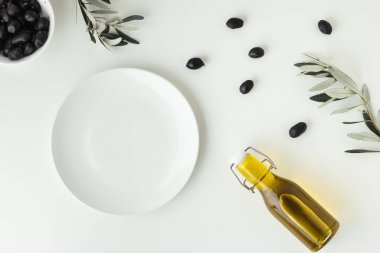 Empty white plate next to white bowl full of black olives and a bottle of olive oil and leaves. White background. Bottle of cold pressed oil. Traditional Greek and Italian food. Flat lay.