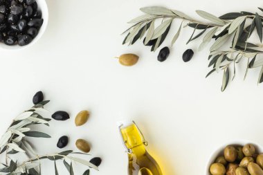Green and black olives in white bowls next to a bottle with olive oil and leaves on a white background.