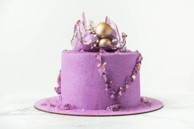Wedding cake with violet cream cheese frosting decorated with caramel vase and golden chocolate spheres on the white background. Luxury anniversary cake with pink chocolate velvet coating