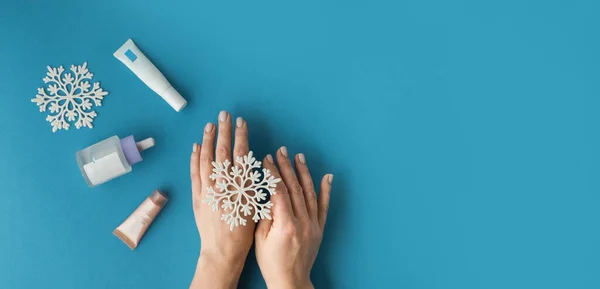 Using moisturizing hand cream during winter season. Female hands smearing white cream on the blue background next to different skin care products. Treatment for the skin advertisement